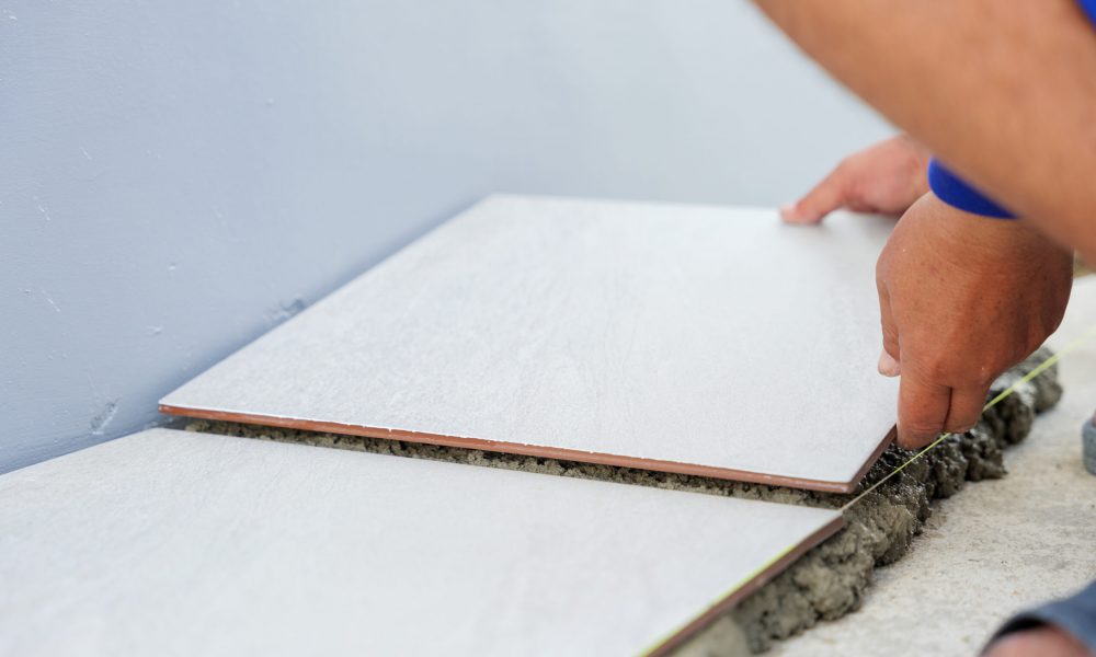 Close-up of a hand placing a white tile onto gray mortar for house flooring. A second tile perfectly aligned beside it demonstrates precision and care in the tiling process.
