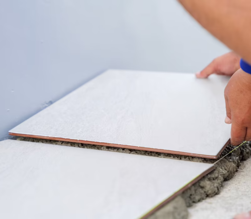 Close-up of a hand placing a white tile onto gray mortar for house flooring. A second tile perfectly aligned beside it demonstrates precision and care in the tiling process.
