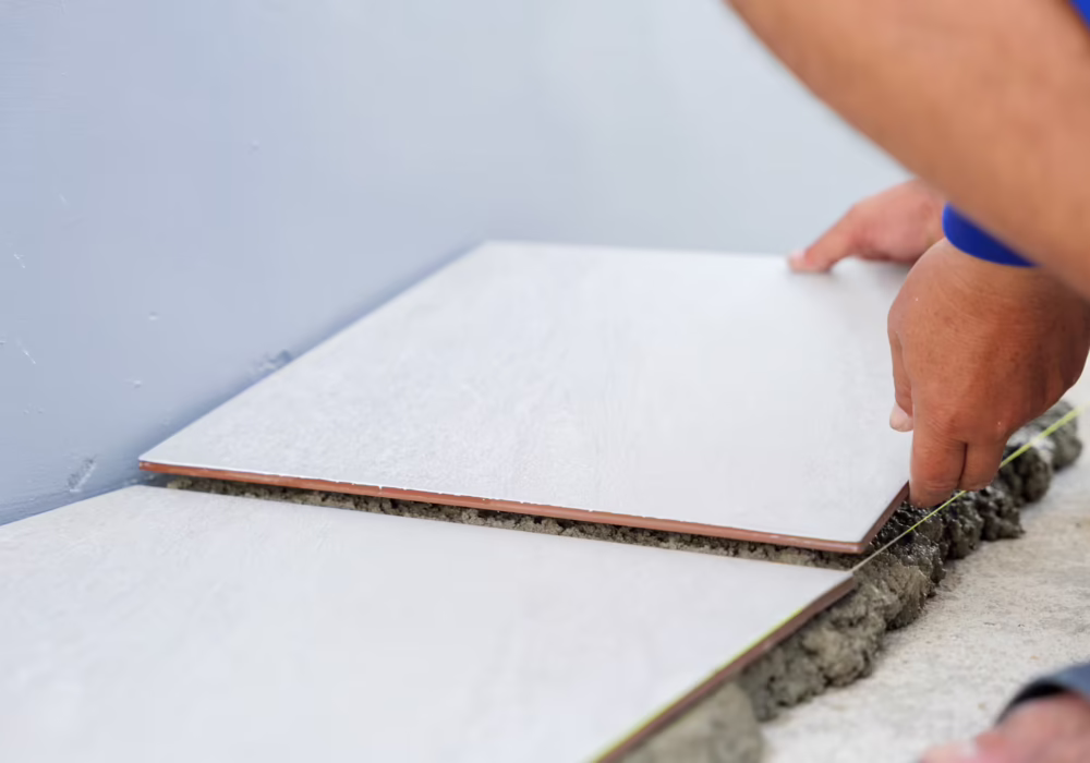 Close-up of a hand placing a white tile onto gray mortar for house flooring. A second tile perfectly aligned beside it demonstrates precision and care in the tiling process.