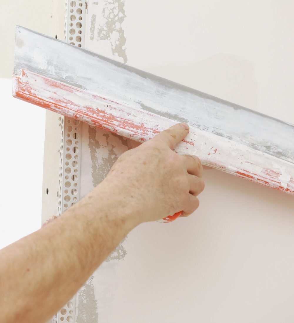 Builder in protective mask using a trowel to add plaster. Plastering wall with putty-knife, close up image. Fixing wall surface and preparation for painting. construction work during quarantine.